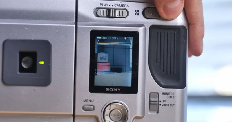 Close-up of a silver Sony camcorder's screen displaying a stacked can and box. A person's hand is pressing a button on the upper right corner of the camera. The camera's buttons and controls are visible.