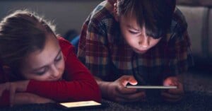 Two children lying on the floor indoors; one child in a plaid shirt is focused on a smartphone, while the other child in red rests their head on their arm next to a phone.