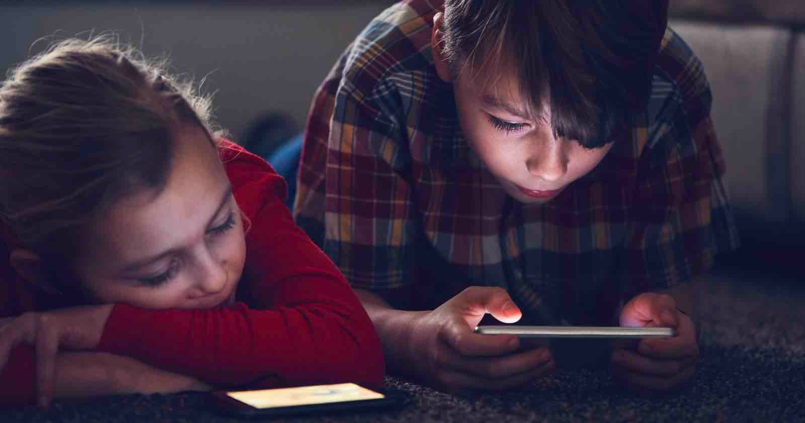 Two children lying on the floor indoors; one child in a plaid shirt is focused on a smartphone, while the other child in red rests their head on their arm next to a phone.