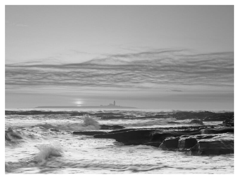 Black and white photo of a rocky coastline with waves crashing, distant island with a tall lighthouse, and dramatic cloudy sky during sunrise or sunset.