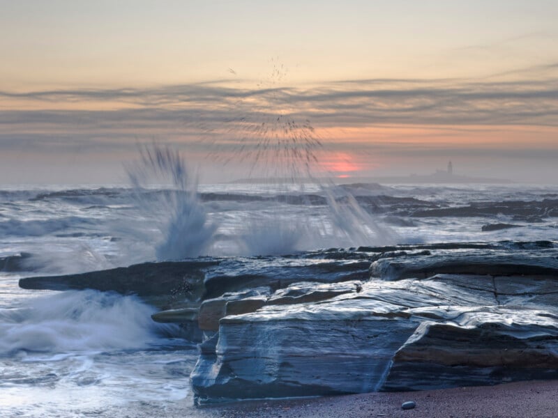 Waves crash against rocky shore at sunset, sending water spray into the air. The sky is filled with soft clouds, and a lighthouse is visible in the distant background through the mist.