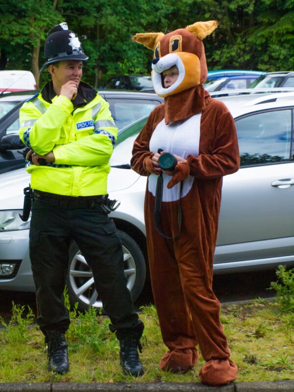 A police officer stands next to a person in a brown squirrel costume holding a dog leash in a parking lot, with cars and greenery in the background.