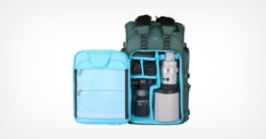 A green camera backpack is open, revealing neatly organized compartments with camera bodies, lenses, and accessories inside a light blue padded interior. The left flap has zipped mesh pockets for additional storage.