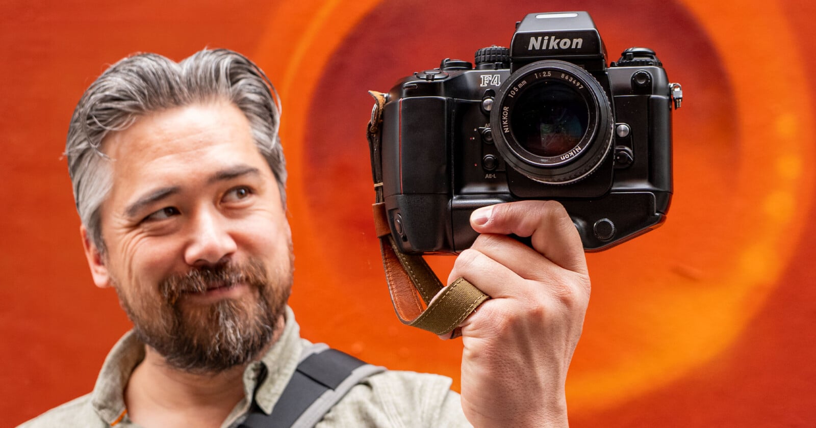 A man with gray hair and a beard smiles while holding a Nikon film camera in front of an orange background with circular patterns.
