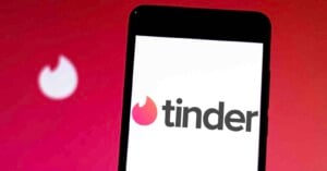 A smartphone displays the Tinder app logo on its screen, with a blurred Tinder flame icon in the background against a red gradient backdrop.
