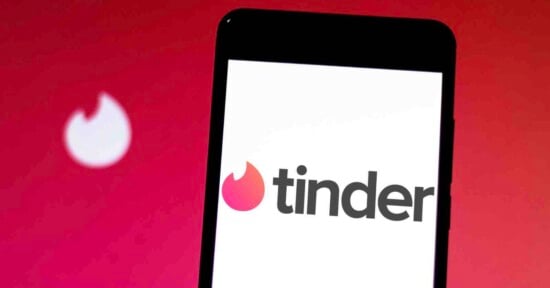 A smartphone displays the Tinder app logo on its screen, with a blurred Tinder flame icon in the background against a red gradient backdrop.