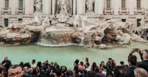 A large crowd of people gathers in front of the Trevi Fountain in Rome, taking photos and admiring the ornate marble statues and turquoise water.