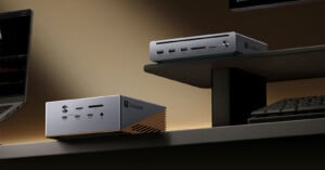 Two compact, silver Thunderbolt-enabled desktop computers sit on a desk and shelf, surrounded by a keyboard, monitor, and laptop in a modern workspace with a warm light.