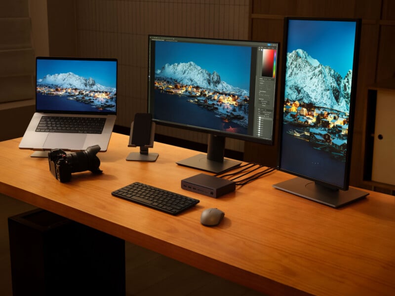 A modern wooden desk with a laptop, two monitors (one vertical), a camera, a keyboard, a mouse, a smartphone stand, and a docking station. All screens display an edited photo of snowy mountains and a town at night.