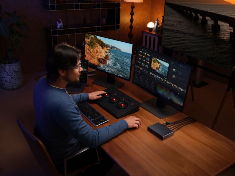 A person sits at a desk using a color grading console with two monitors displaying video editing software and scenic beach footage. The workspace is warmly lit and organized, with a keyboard and other equipment on the desk.