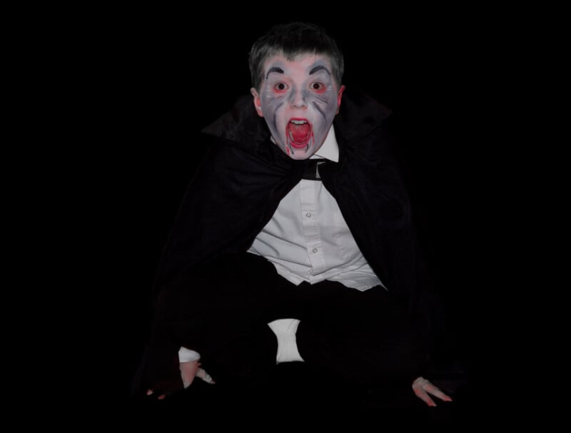 A child dressed as a vampire crouches, wearing a black cape, white shirt, and makeup with a pale face, dark eyes, and red lips, mimicking a scream or roar against a black background.