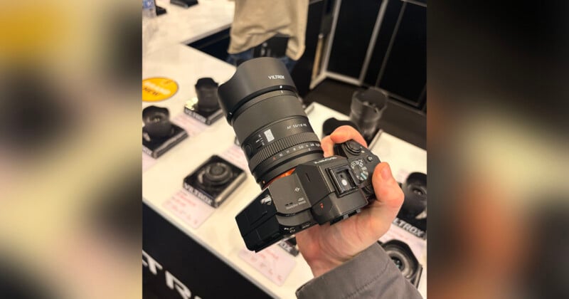A person holds a Sony mirrorless camera with a large Viltrox lens, above a display table featuring several other camera lenses and accessories.