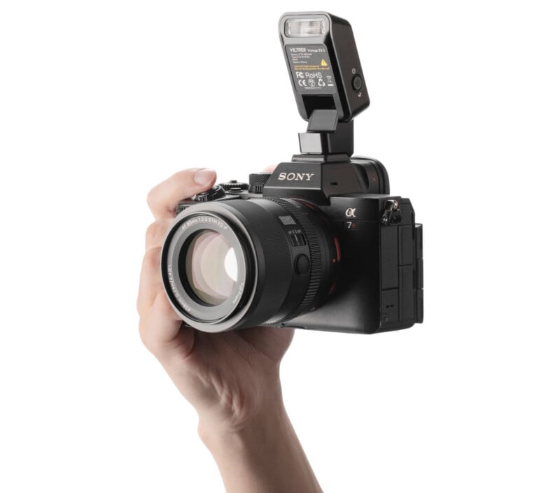 A hand holding a Sony mirrorless camera with a large lens and an external flash attached, isolated on a white background.
