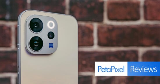 Close-up of a smartphone's rear camera module with three lenses, set against a blurred brick wall. At the bottom right, the Petapixel Reviews logo appears.