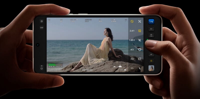 A person holds a smartphone horizontally, filming a woman in a yellow dress sitting on rocks by the sea, with camera settings visible on the screen.