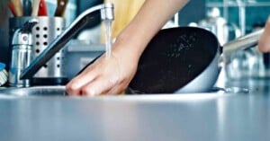 A person’s hand washes a black pan under running water in a kitchen sink, with utensils and a faucet visible in the background.