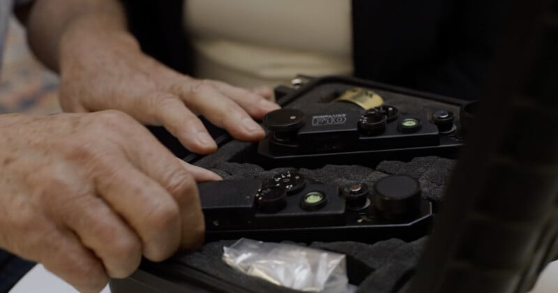 A close-up of two hands opening a foam-lined case containing black electronic devices with dials, switches, and bubble levels, along with a small plastic bag.