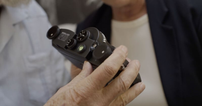A close-up of an older person's hand holding a vintage black film camera, with two people partially visible in the background.