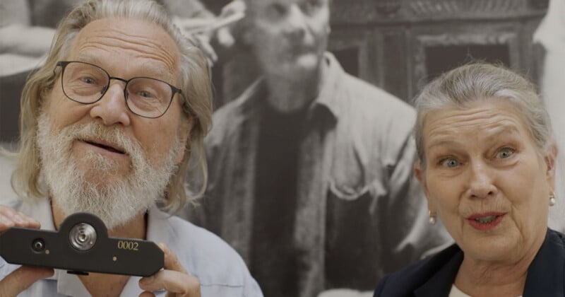 An older man with glasses and a white beard smiles while holding a camera device, standing next to an older woman with gray hair, both in front of a black-and-white photo background.