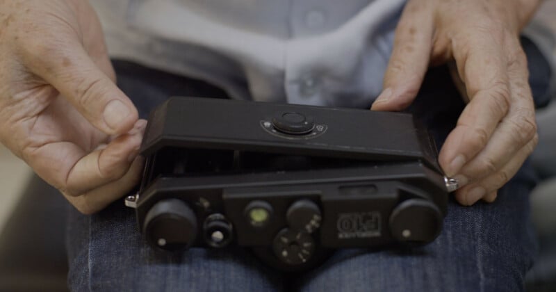 Close-up of an elderly person’s hands holding a vintage black camera device, resting on their jeans-clad lap. The camera appears to be partially open or being examined.