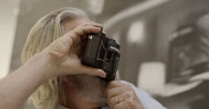 A person with long gray hair and a beard holds a WIDELUX camera up to their face, preparing to take a photo. The background is blurred and features monochrome artwork.