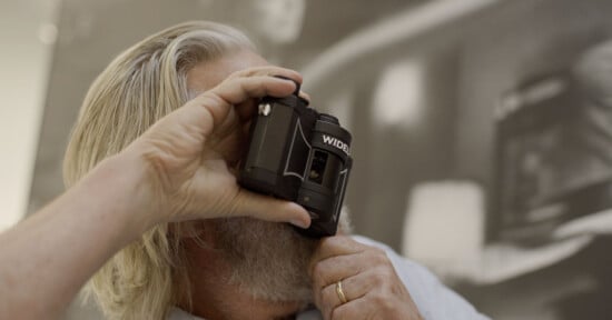 A person with long gray hair and a beard holds a WIDELUX camera up to their face, preparing to take a photo. The background is blurred and features monochrome artwork.