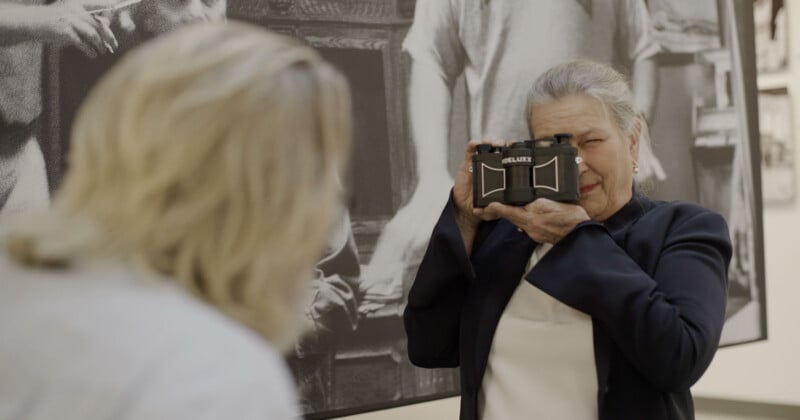 An older woman in a dark blazer playfully holds a vintage camera up to her face, aiming it at another person with light hair, in front of a large black and white photo in an art gallery.
