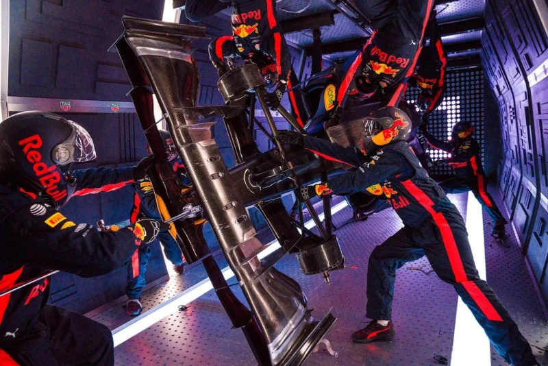 A Red Bull Racing pit crew performs a simulated pit stop in a zero-gravity environment, working on a Formula 1 car part while floating, all wearing matching team uniforms and helmets.
