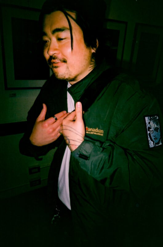 A man with a mustache and beard stands indoors wearing a dark green jacket with "Canadian" on it and an anime patch on the sleeve. He has black hair, rings, and is holding his jacket open slightly with one hand on his chest.
