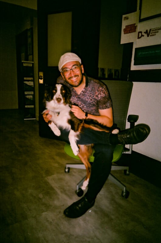 A smiling man wearing glasses and a cap sits on an office chair, holding a relaxed black, brown, and white dog in his lap. They are indoors, with framed posters on the wall in the background.