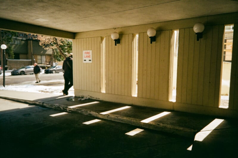 A covered parking area with vertical slats letting sunlight stream through, casting shadows on the ground. A sign reads "FIRE LANE." Two people walk outside on a snowy sidewalk near parked cars.