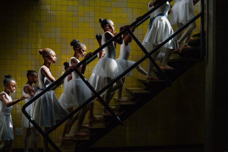 A group of young ballet dancers in white tutus ascend a staircase with yellow tiled walls, bathed in dramatic light and shadow.