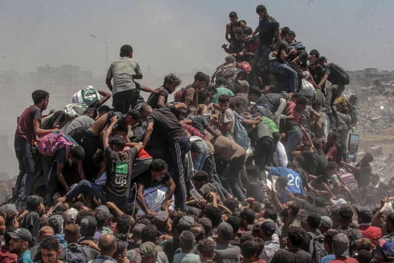 A large crowd of people, mostly men and boys, climb and gather on a mound of earth amid a dusty, chaotic environment, suggesting urgency or desperation. Many appear to be reaching upwards or helping each other.