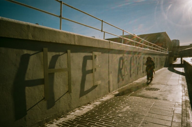 A person walks along a sunlit sidewalk next to a concrete wall with large shadowed letters and railings on top. The scene is bright, with strong sunlight casting long shadows.