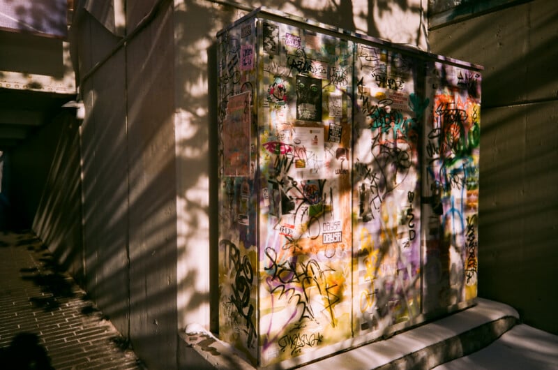 A utility box covered in colorful graffiti and stickers stands against a concrete wall, with tree shadows cast across it in bright sunlight.