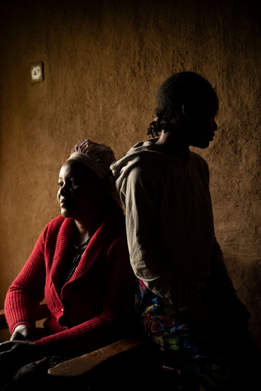 A woman in a red sweater sits beside a girl standing in a hoodie, both in a dimly lit room with brown walls. The woman faces the light, while the girl is in shadow, their expressions calm and contemplative.