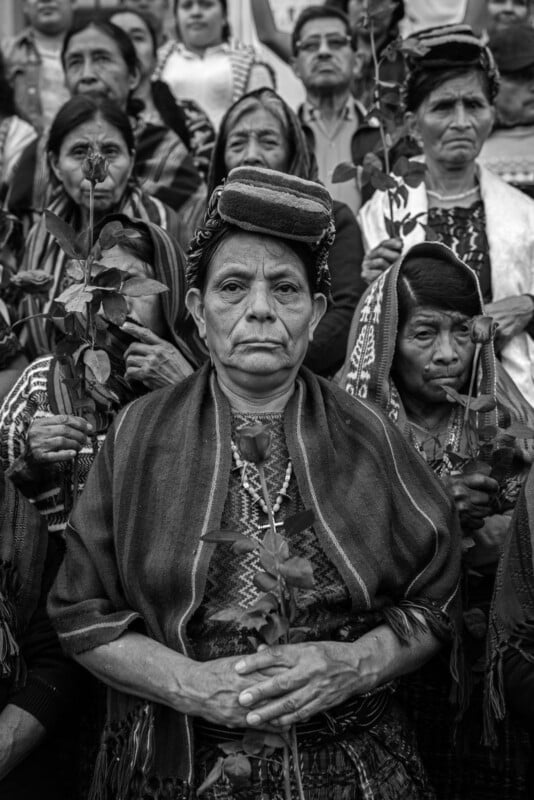 A group of Indigenous women in traditional clothing hold roses and look solemnly at the camera; the central woman sits in front with a woven cloth on her head. The photo is black and white.