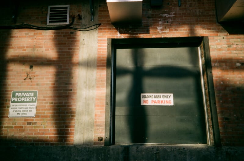 A loading bay door in a brick wall has a sign reading "Loading Area Only No Parking." Nearby, a "Private Property" sign warns unauthorized vehicles will be towed. Shadows fall across the wall and door.