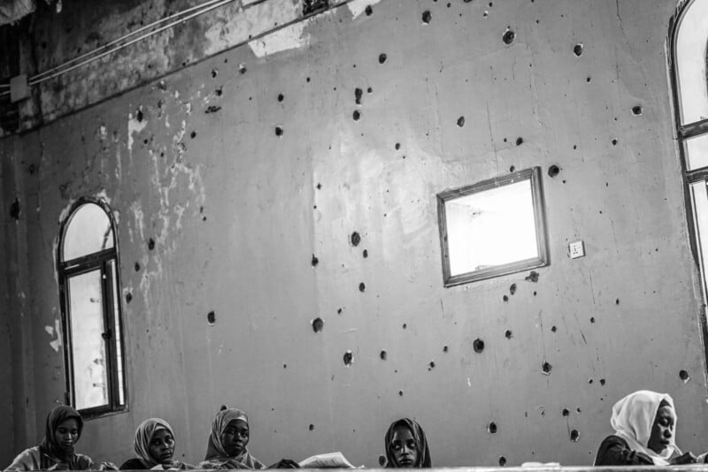 Five women wearing headscarves sit beneath a wall marked with bullet holes in a rundown room with arched and rectangular windows. The atmosphere appears somber and tense.