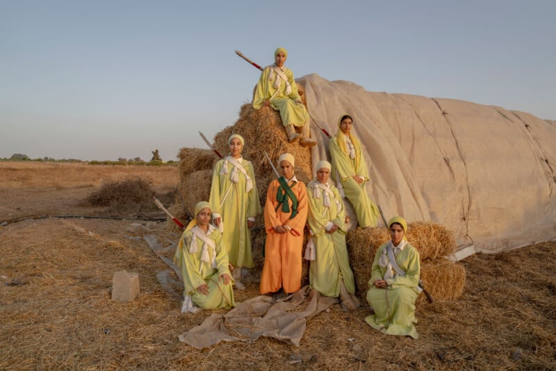 Seven women wearing long pastel dresses and headscarves pose on and around stacked hay bales in an open field at sunset; a large tarp covers part of the hay. The sky is clear and the landscape is dry.