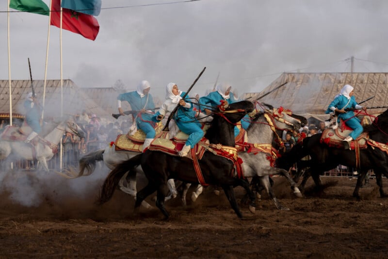 Women dressed in blue and white traditional clothing ride decorated horses through smoke during a cultural equestrian performance, with flags and a crowd visible in the background.
