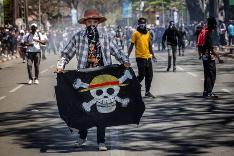 A person in a straw hat and mask holds a black flag with a skull and crossed bones, resembling the One Piece anime logo, during a street protest with others in the background.