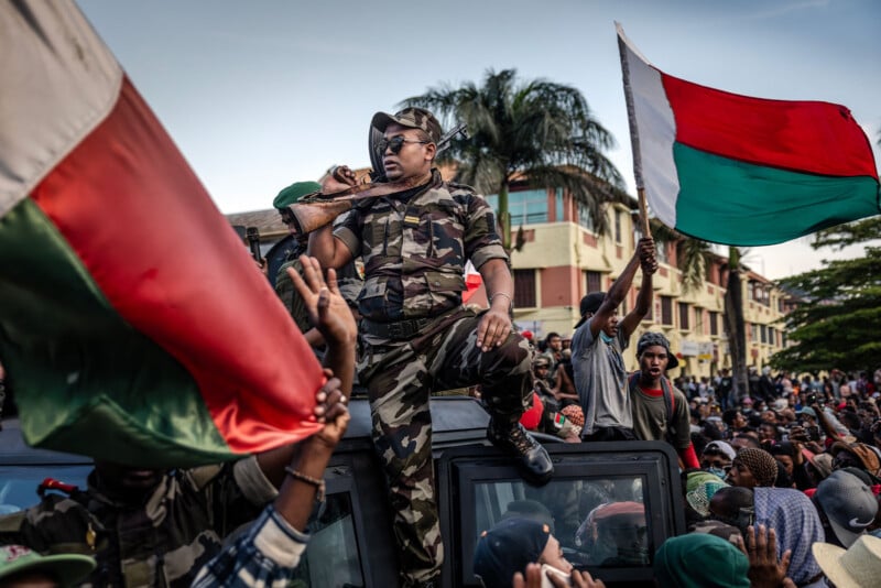 A soldier in camouflage sits on a vehicle, speaking into a radio, surrounded by a crowd waving large red, green, and white flags in an urban setting with palm trees and buildings in the background.