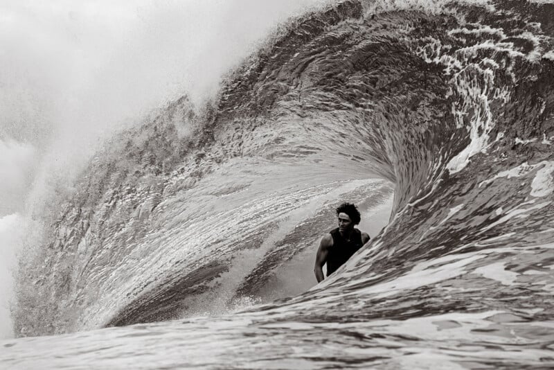 A surfer rides inside the barrel of a powerful, curling wave. Water arcs dramatically overhead, framing the surfer in the tube. The image is in black and white, emphasizing the wave’s texture and motion.