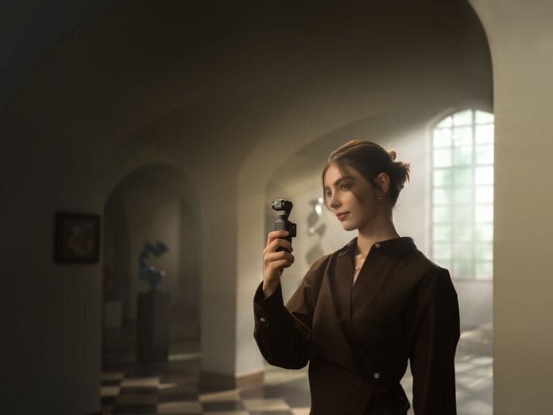 A woman in a brown dress stands in a sunlit room with arched hallways, holding a small handheld camera or gimbal and looking at it thoughtfully. Soft light streams through a large window.