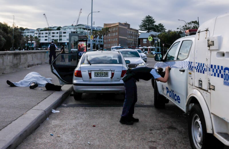 A police officer leans on a police vehicle near a silver car, with a person lying on the ground next to the car. Emergency vehicles and buildings are visible in the background on a city street.