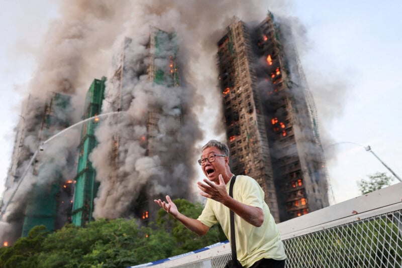 A distressed man gestures urgently in the foreground as two high-rise buildings behind him are engulfed in flames and thick smoke, with firefighters spraying water from hoses toward the burning towers.