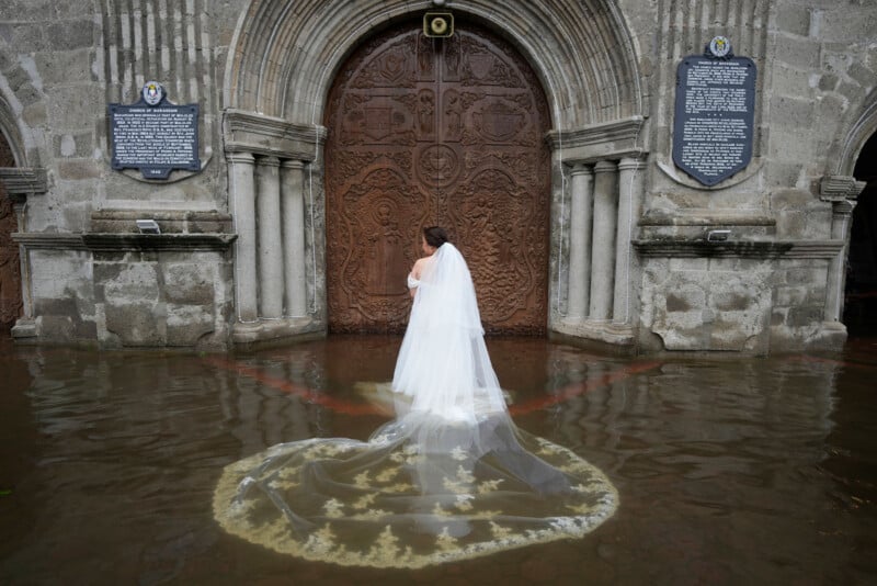 A bride in a white wedding dress and veil stands in ankle-deep floodwater outside large ornate church doors, with her back to the camera and her dress trailing in the water.