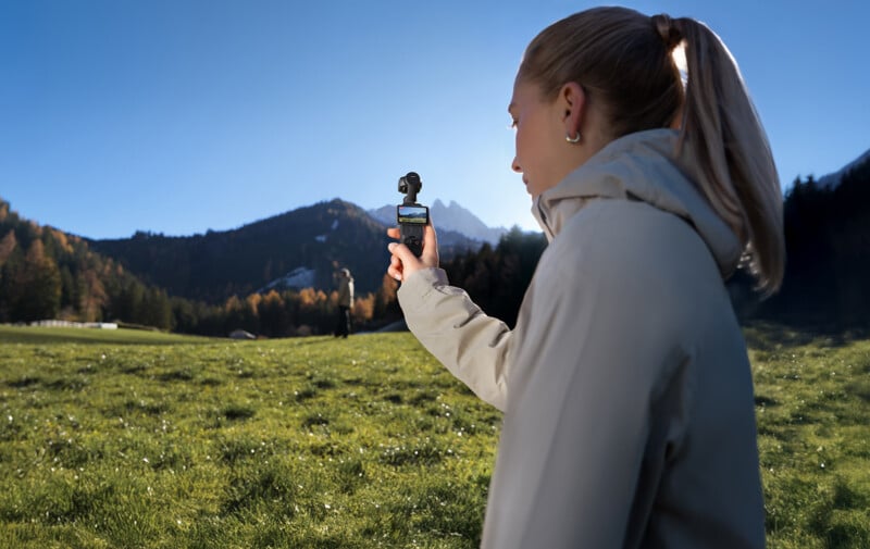 A person with blond hair in a ponytail, wearing a light jacket, stands in a grassy field taking a video with a small handheld camera. Forested hills and mountains appear in the background under a clear blue sky.