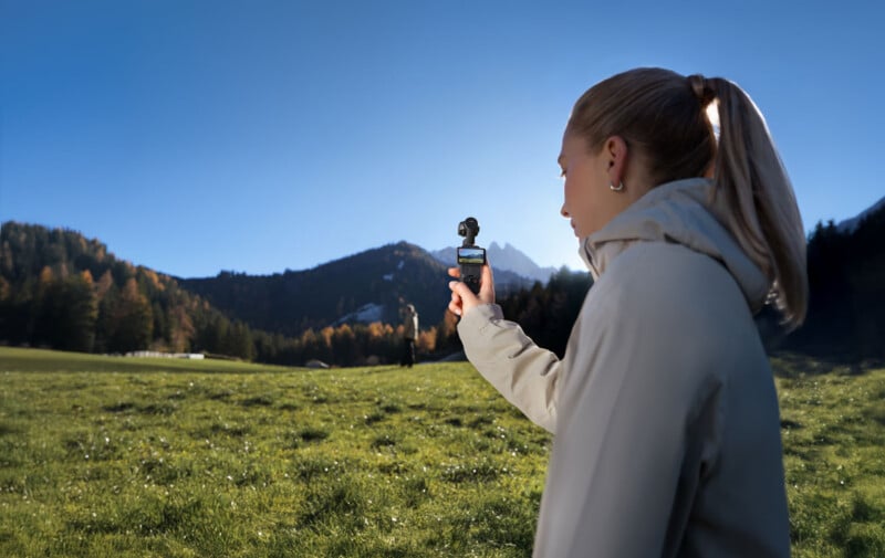 A woman in a light jacket stands in a grassy field with mountains in the background, holding a small camera or gimbal, facing the landscape on a sunny day.